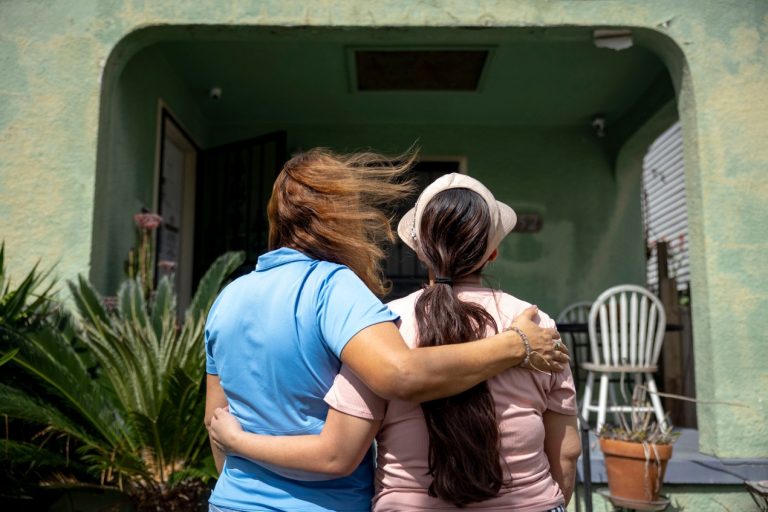 Louisiana’s Reporting Law Chills Immigrant Medicaid Applications Two women stand side-by-side, each with an arm around the other, and face away from the camera toward a building.