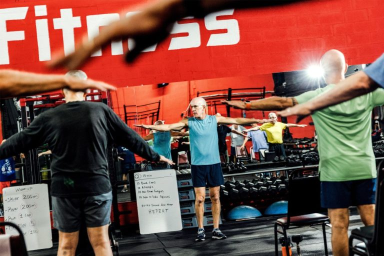 Larry Gruber stands in front of a mirror at a gym, his arms extended on both sides, stretching. Blurred arms in the foreground show students copying his move.