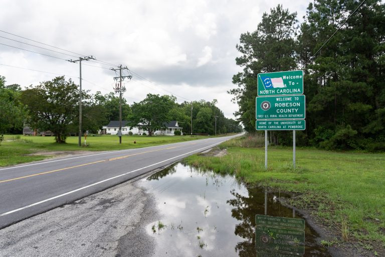 A photo taken from the side of the road shows three signs. They read: "Welcome to North Carolina." "Welcome to Robeson County; first U.S. rural health department." "Home of the University of North Carolina at Pembroke."