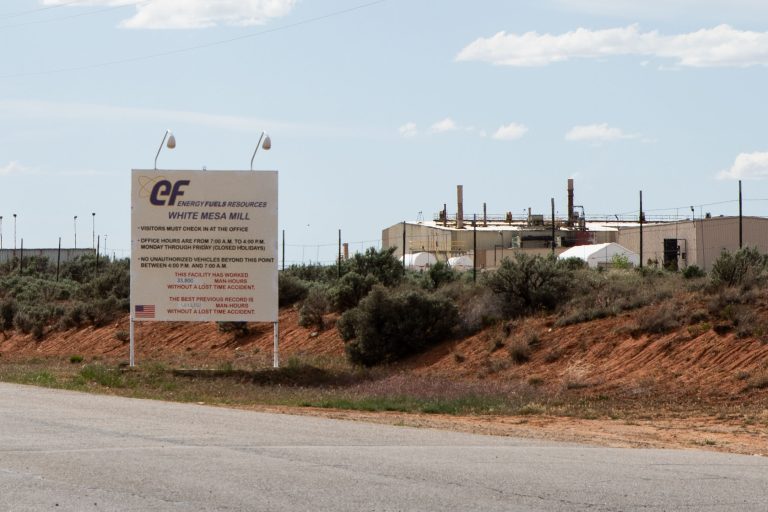A photo shows a sign in front of White Mesa Mill. The facility is seen in the background.